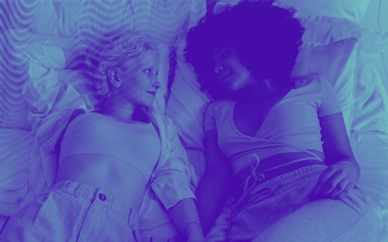 Duas mulheres deitadas juntas em uma cama, em um cenário roxo relaxante, refletindo um momento de intimidade e amizade