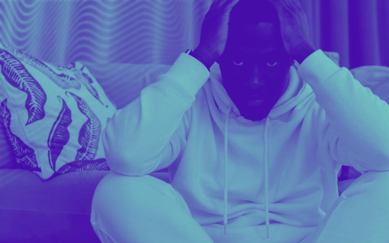 Homem sentado à frente do sofá com as mãos na cabeça, alusivamente de pensativo